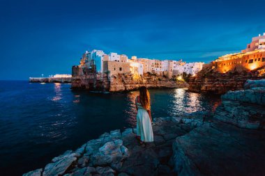 Polignano a Mare, İtalya - Ağustos 2021: Apulian köyü Polignano a Mare 'nin önünde kayalıklarda genç bir kadın gün batımında (mavi saat)