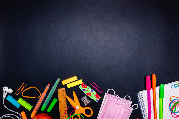Milan, Italy - January 2022: Series of stationery items, colored pens, labels, paper clips, calculator, scissors, pink face masks and more positioned below on black background, back to school concept, flat lay, blank space