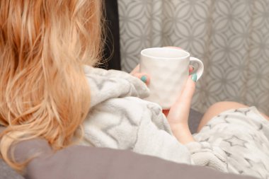 Kanepede oturan bir kız banyodan sonra beyaz bardaktan lezzetli bitki çayı içer. Samimi bir fotoğraf. Kadın rahat bir yaşam tarzıyla ev koltuğunda eğleniyor..