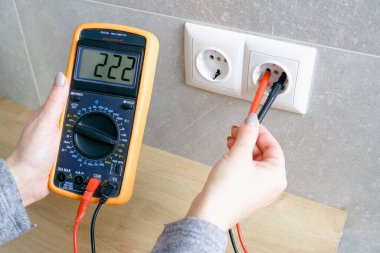 Çoklu metresi olan bir kadın elektrik şebekesinin voltajını kontrol ediyor. Ellerin yakın görüntüsü ve soketin yanındaki alet..