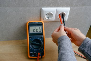 Çoklu metresi olan bir kadın elektrik şebekesinin voltajını kontrol ediyor. Ellerin yakın görüntüsü ve soketin yanındaki alet..