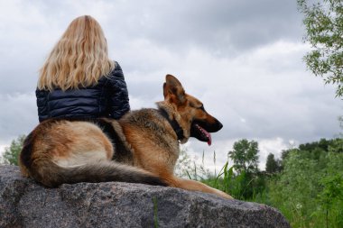 Alman çoban köpeği olan genç bir kadın bulutlu bir bahar gününde gölün kenarında büyük bir granit taşın üzerinde oturur..