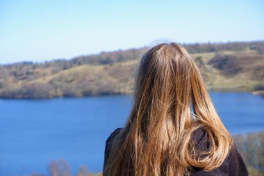 A girl is resting on a clear day on the shore of a blue lake with wooded hilly shores in early spring. Secluded relaxation concept. Shooting back without face