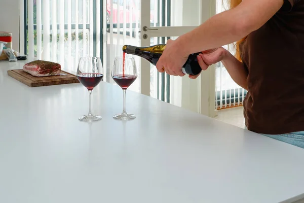 A woman pours red wine from a bottle into wine glasses. Modern white kitchen clean interior design. Glass of wine in female hand.