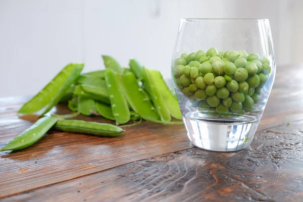 Green peas in glass bowl. fresh pea in the pod with green leaves. green peas on a brown wodden table. Shelled peas in a glass with the pods on the side