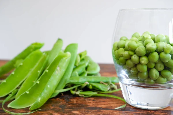 Green peas in glass bowl. fresh pea in the pod with green leaves. green peas on a brown wodden table. Shelled peas in a glass with the pods on the side