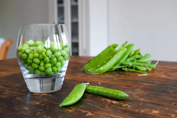 Green peas in glass bowl. fresh pea in the pod with green leaves. green peas on a brown wodden table. Shelled peas in a glass with the pods on the side