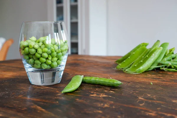 Green peas in glass bowl. fresh pea in the pod with green leaves. green peas on a brown wodden table. Shelled peas in a glass with the pods on the side