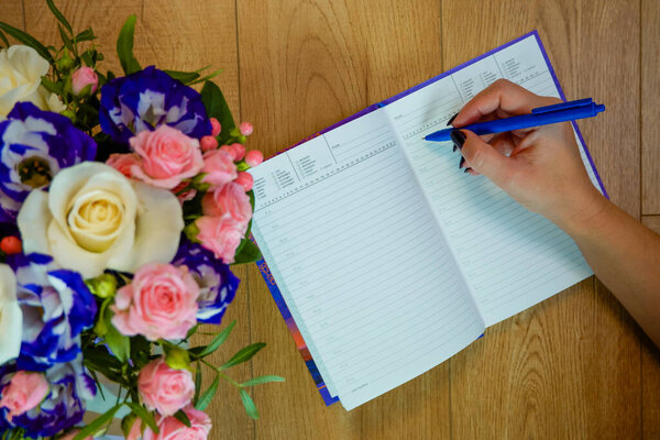 Female hands writing in open notebook and bouquet of roses on old wooden table. Top view. Hand writing a love note in a notebook.