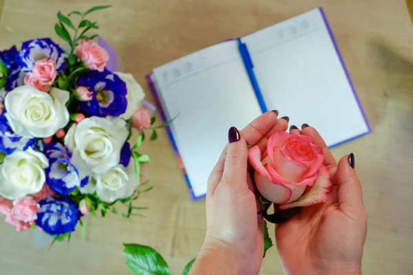 Female hands hold a rose flower. Notebook, diary with a pen on a wooden table near a bouquet of multi-colored roses in the background. Top view, flat lay.