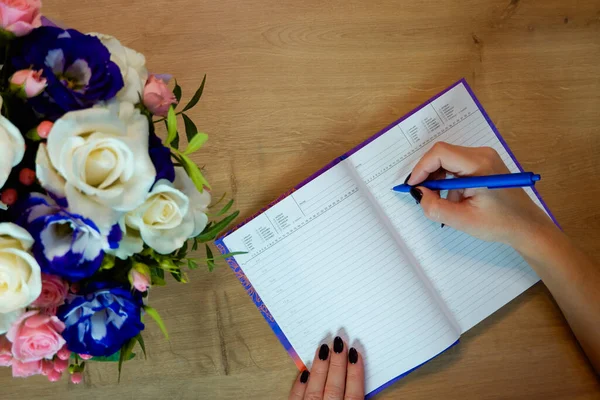 Female hands writing in open notebook and bouquet of roses on old wooden table. Top view. Hand writing a love note in a notebook.