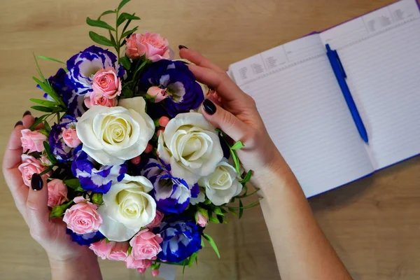 Female hands hold a rose flower. Notebook, diary with a pen on a wooden table near a bouquet of multi-colored roses.