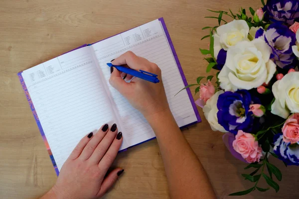 Female hands writing in open notebook and bouquet of roses on old wooden table. Top view. Hand writing a love note in a notebook.