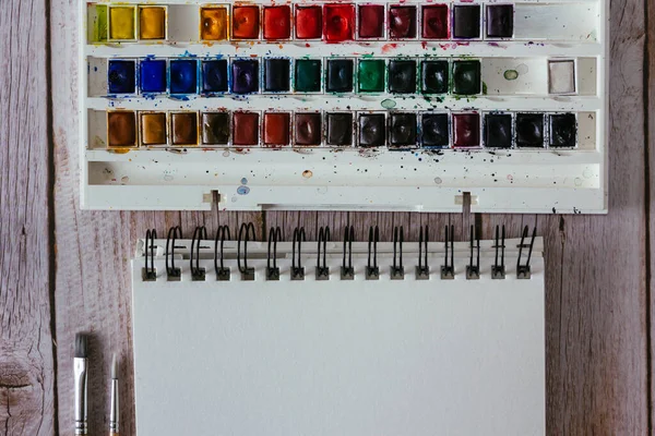 Bir palet suluboya boya, boş kağıt ve ahşap arka planda fırçalar. Sanatçının çalışma alanı. Uzayı kopyala Seçici odak.