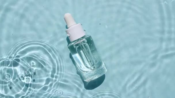 Стеклянная косметическая бутылка с пипеткой лежит на поверхности воды. Вид сверху на воду синего цвета. Чистая голубая вода с отражением солнечного света. Пример упаковки для дизайна, рекламы.4k — стоковое видео
