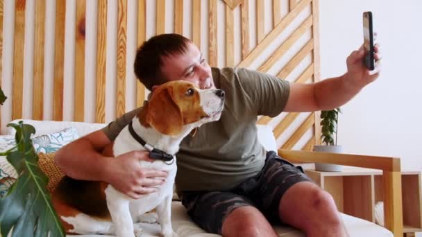 Ein fröhlicher junger Mann sitzt am Tisch und umarmt ihren Hund Beagle beim Videoanruf. Der Besitzer des Hundes macht ein Selfie mit dem Haustier per Handy. Zeitlupe. — Stockvideo