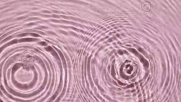 Вид сверху медленное движение капли падает в воду и расходящиеся круги воды на розовом фоне. Капля воды синего цвета. Чистая вода с отражениями, преломление солнечного света.4k — стоковое видео