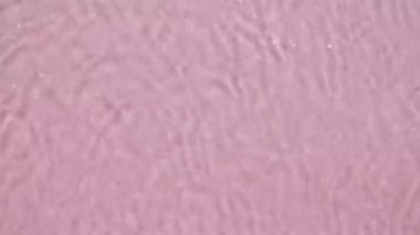 Su yüzeyi üst görünümü. Su pembe renkte sıçrıyor. Saf mavi su, güneş ışığını yavaş çekimde yansıtıyor. Hareket temiz yüzme havuzu dalgalanmaları ve el sallama. 4k