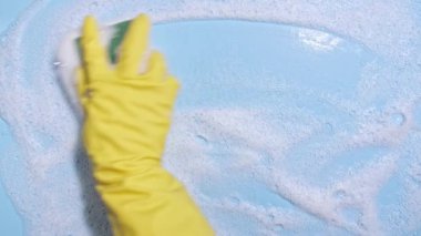 Sarı lastik bir eldiven, temiz bir sünger tutar ve mavi arka planda sabunlu bir köpüğü siler. Temizlik konsepti, temizlik hizmeti. Pankart. Düz yerde, üst tarafta. Yüksek kalite 4k görüntü