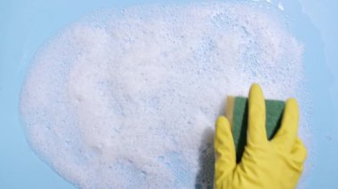 Sarı lastik bir eldiven, temiz bir sünger tutar ve mavi arka planda sabunlu bir köpüğü siler. Temizlik konsepti, temizlik hizmeti. Pankart. Düz yatış, üst görünüm