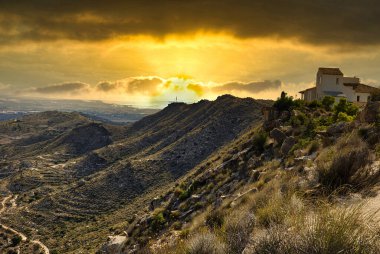 Dağların ve evin üzerinde gün batımı, dağın tepesinde, saklanan güneşin turuncu ışığıyla aydınlanıyor. İspanya, Alicante vilayeti. Yatay görüş..