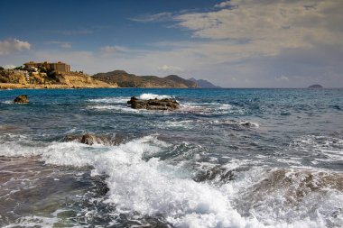 Alicante eyaletinin Altea kentindeki körfezin kayalık kıyılarına dalgalar çarpıyor. Mavi gökyüzünün altında beyaz bulutlu yatay manzara