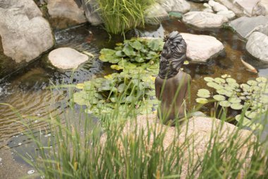 Parktaki gölet. Taşlar ve suyla suni manzara. Çelik bir denizkızı menekşelerin arasında bir kayanın üzerinde oturur..