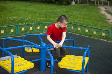 Atlıkarıncadaki çocuk. Çocuk oyun parkındaki çocuklar için atlıkarıncada dönüyor. Ortaokul çocuğu tek başına oynuyor. Kısa saçlı, kırmızı tişörtlü bir çocuk. Adam tırabzana sıkıca tutunuyor..
