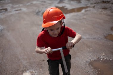 Motosikletli turuncu kasklı çocuk. Yazın sokakta gezen bir çocuk. Kasklı çocuk toprak yolda gidiyor. Oğlumun geri kalanı yaz tatilinde. Küçük okul çocuğu bahçede tek başına oynuyor..