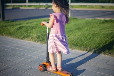 Çocuk scooterlı bir kız. Parkta çocuk gezintisi. Kız gitmek için zorluyor. Yazın hamile. Anaokulu öğrencisi karede yalnız. Şehirde yaz günü.