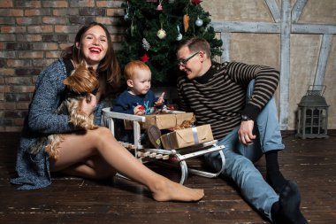 Küçük kızıl saçlı bir çocuk, Noel ağacının yanındaki ahşap kızağın üzerinde oturuyor. Yanında da anne ve babası ve köpeği var. Noel ağacının yanında köpekli mutlu bir aile.