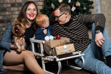 Küçük kızıl saçlı bir çocuk, Noel ağacının yanındaki ahşap kızağın üzerinde oturuyor.