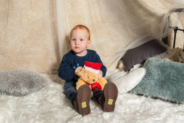 Kızıl saçlı küçük çocuk bir Noel ayısıyla oynuyor. Küçük çocuk en sevdiği oyuncağına sarılıyor. Kırmızı Noel şapkalı bir ayı. Çocuklar içeride oynuyor.