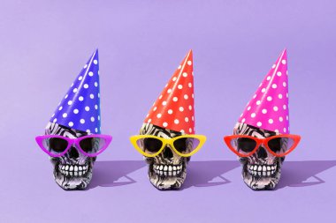 Three Halloween skulls with colorful sunglasses and polka dot party hats against purple background. Creative creepy autumn holiday concept. Trendy vibrant colors and sunny day shadows.