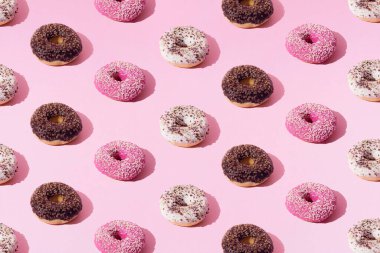 Beyaz, pembe ve çikolata jöleli halka çörekler. Pembe pastel arka planda renkli kırıntılar. Desen.