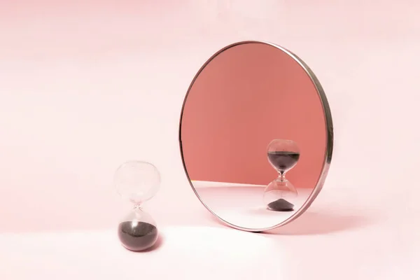 Sand clock stands in front of mirror. Pink background