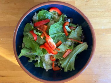 Taze marul yaprakları, çıtır çıtır kırmızı biber, salatalık, otlar, domates ve taze soğan, mavi bir seramik kasede vejetaryen ve vejetaryen salatası, ahşap mutfak masasının yüzeyinde toprak kaplamaları düz bir görünüm.