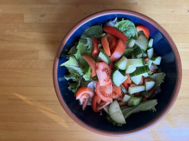 Taze marul yaprakları, çıtır çıtır kırmızı biber, salatalık, otlar, domates ve taze soğan, mavi bir seramik kasede vejetaryen ve vejetaryen salatası, ahşap mutfak masasının yüzeyinde toprak kaplamaları düz bir görünüm.