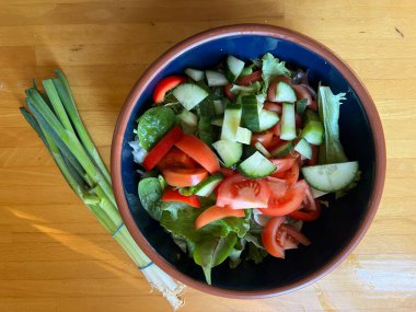 Taze marul yaprakları, çıtır çıtır kırmızı biber, salatalık, otlar, domates ve taze soğan, mavi bir seramik kasede vejetaryen ve vejetaryen salatası, ahşap mutfak masasının yüzeyinde toprak kaplamaları düz bir görünüm.