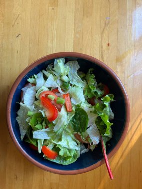 Taze marul yaprakları, çıtır çıtır kırmızı biber, salatalık, otlar, domates ve taze soğan, mavi bir seramik kasede vejetaryen ve vejetaryen salatası, ahşap mutfak masasının yüzeyinde toprak kaplamaları düz bir görünüm.