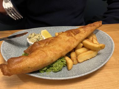 Balık ve patates kızartması, günün taze morina balığı, kızarmış bira hamuru, yumuşak merkezli patates cipsi ve deniz kenarındaki deniz ürünleri restoranında mavi tabakta limon dilimi.