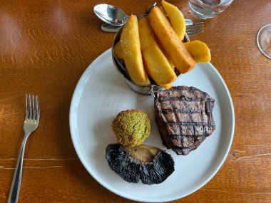 Biftek menüsünü az pişmiş ızgara etle kapayın. Baharatlı cipsler teneke kutuda ve mantarlar beyaz tabak ahşap masa zemininde. Şarap kadehi ve çatal bıçak seti de restoranın manzarasında.