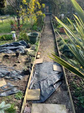 Bahçe tasarımı projesinin peyzajı düz bir yol inşa ediyor. Zemin sert temeller için kazılmış, ahşap kenarlar ve antika tuğla yüzey için döşenmiş siyah ışık bloklama örtüsü için hazır.