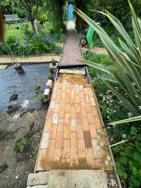 Geri kazanılmış kil, sarı ve kırmızı antika ev taşlarından oluşan tuğla yol inşaatına yakın. Bahçenin düz bir yolunu inşa edin. Çimlerin ve gül kemerinin altında bitkiler ve çevreleyen yeşillik desenli desenler oluşturun.