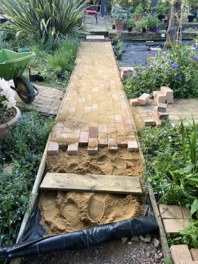 Geri kazanılmış kil, sarı ve kırmızı antika ev taşlarından oluşan tuğla yol inşaatına yakın. Bahçenin düz bir yolunu inşa edin. Çimlerin ve gül kemerinin altında bitkiler ve çevreleyen yeşillik desenli desenler oluşturun.