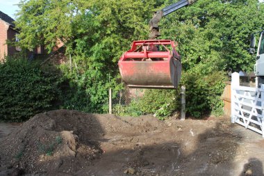Close up  soil grab digger heavy motor machinery with digging arm bucket moving earth, lifting to clear concrete drive and dispose of rubble in re-landscaping organic garden to create level ground 