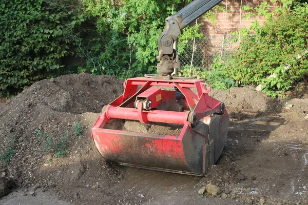 Close up  soil grab digger heavy motor machinery with digging arm bucket moving earth, lifting to clear concrete drive and dispose of rubble in re-landscaping organic garden to create level ground