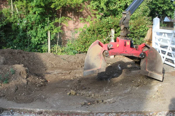 Close up  soil grab digger heavy motor machinery with digging arm bucket moving earth, lifting to clear concrete drive and dispose of rubble in re-landscaping organic garden to create level ground