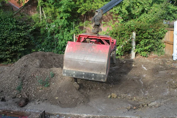 Close up  soil grab digger heavy motor machinery with digging arm bucket moving earth, lifting to clear concrete drive and dispose of rubble in re-landscaping organic garden to create level ground