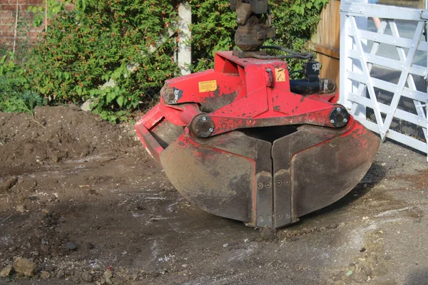Close up  soil grab digger heavy motor machinery with digging arm bucket moving earth, lifting to clear concrete drive and dispose of rubble to create level ground re-landscaping organic garden drive with white gate